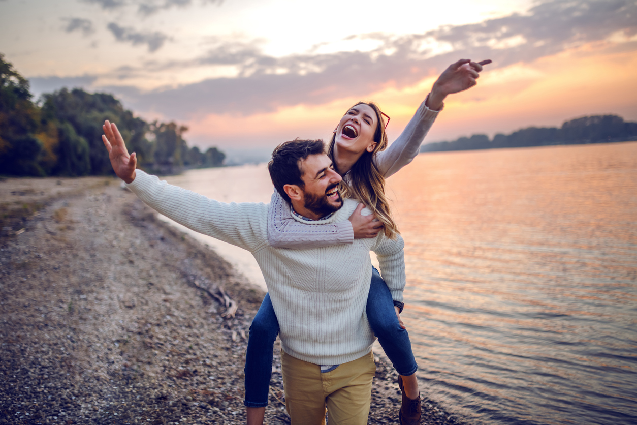 Playful caucasian couple having piggyback on coast next to river. In background is sunset.