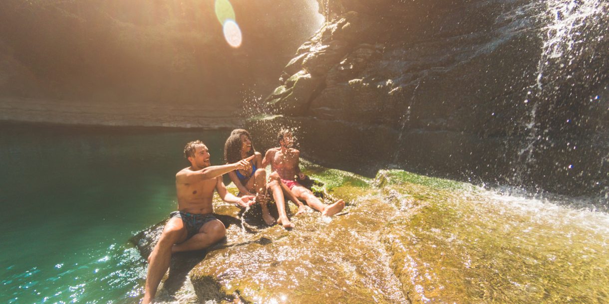 Group of friends having fun under waterfalls river – Young people swimming inside emerald water lagoon – Travel, summer and friendship concept – Focus on left guy face