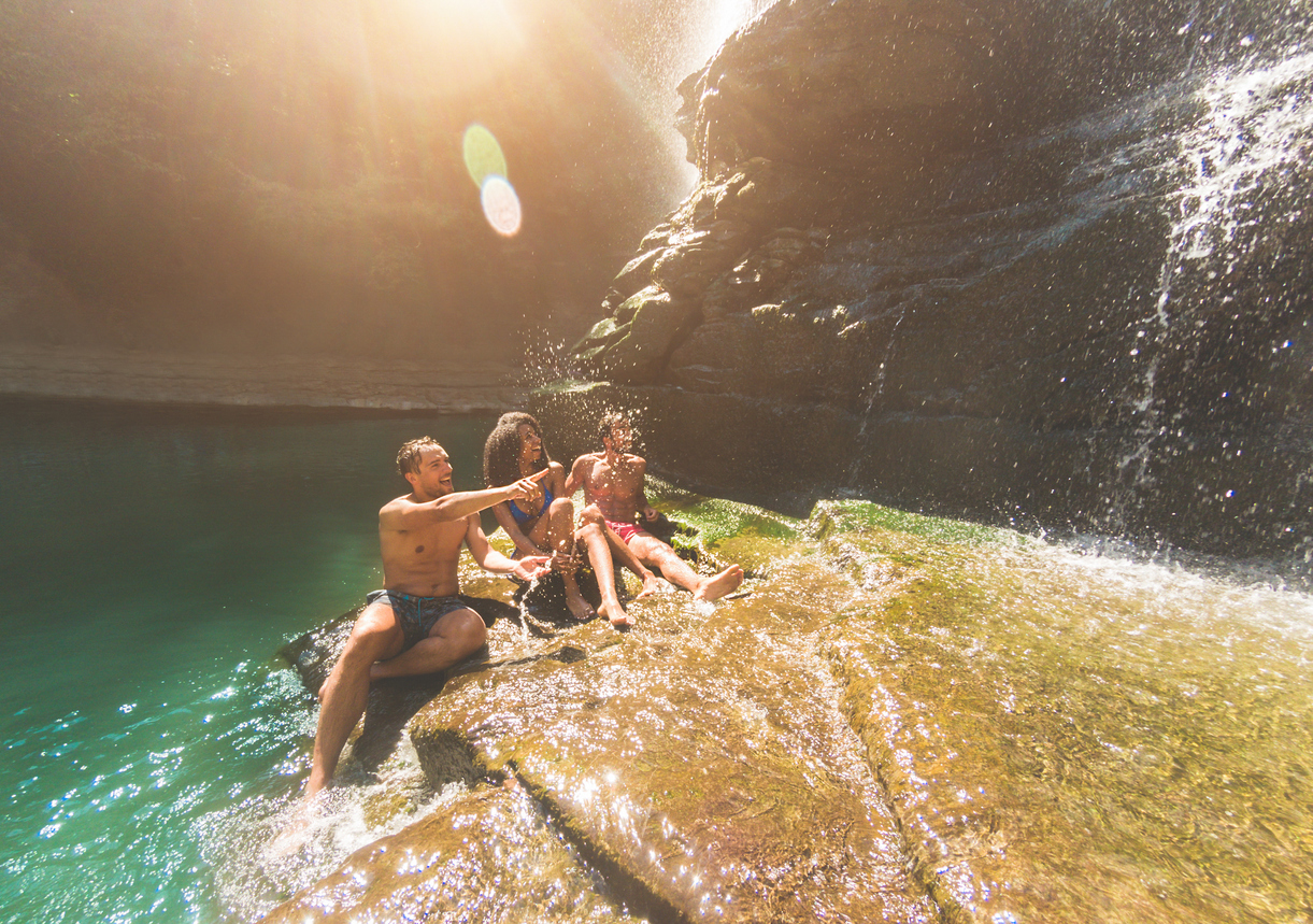 Group of friends having fun under waterfalls river – Young people swimming inside emerald water lagoon – Travel, summer and friendship concept – Focus on left guy face