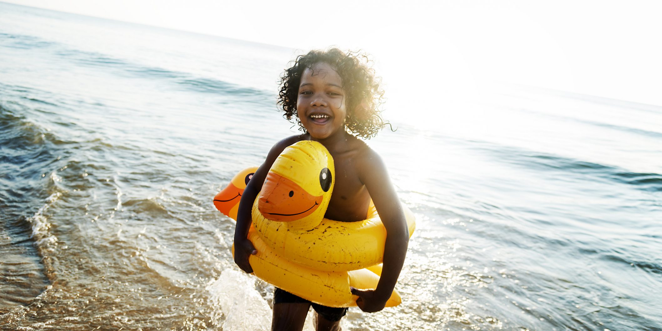9-praias-para-levar-as-criancas-em-Santa-Catarina Criança sorrindo usando uma bóia de pato amarelo saindo do mar na praia