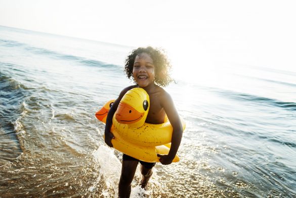 Criança sorrindo usando uma bóia de pato amarelo saindo do mar na praia