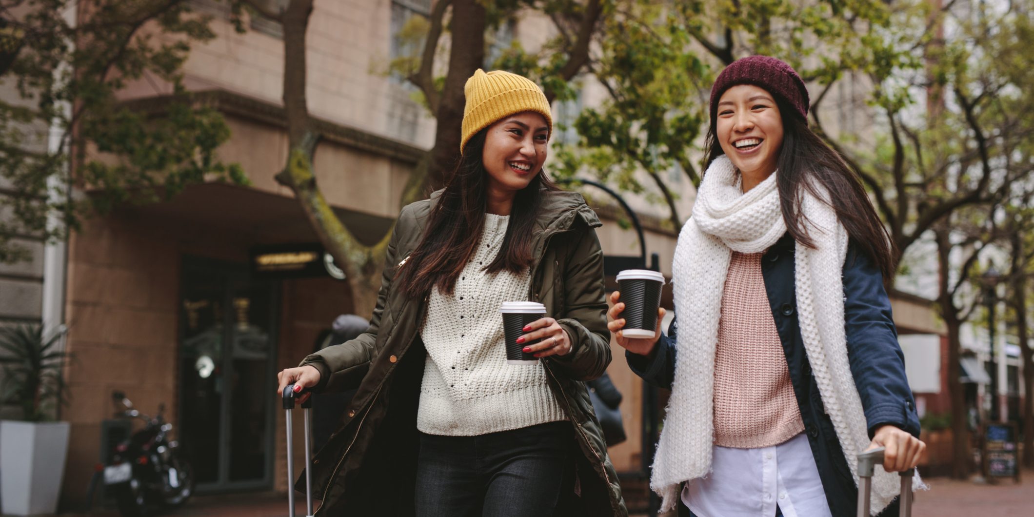 Imagem de duas turistas sorrindo e tomando um café, andando com suas malas de mão.